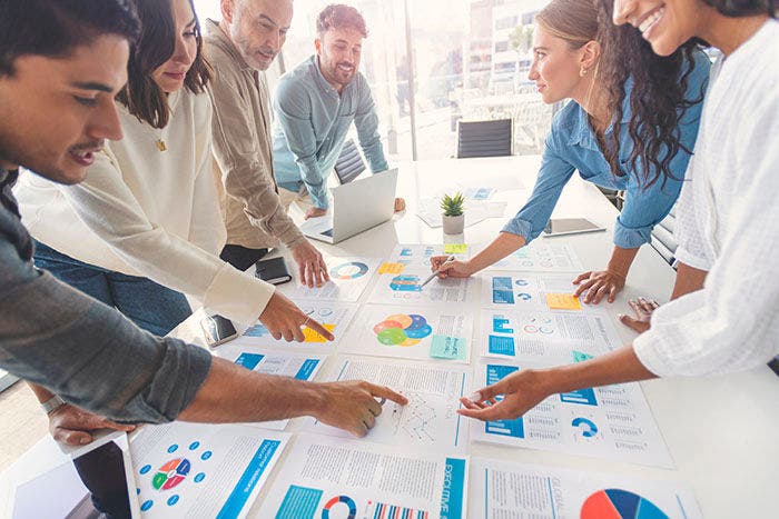 Multi racial group of people working with Paperwork on a board room table at a business presentation or seminar. The documents have financial or marketing figures, graphs and charts on them. People are pointing to different documents. There is a laptop on the table